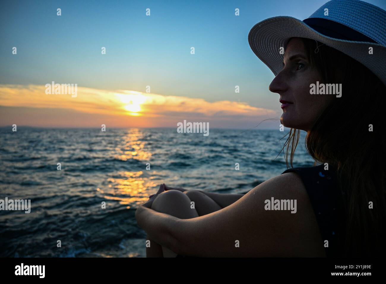 Una bella donna che indossa un abito estivo e un cappello da sole posa al tramonto sulla spiaggia. Foto Stock