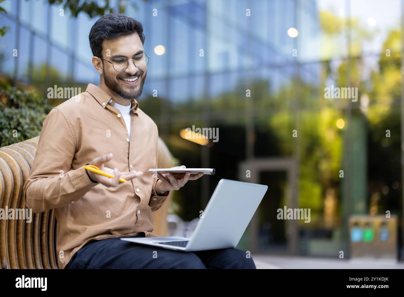 Un giovane sorridente utilizza notebook e tablet per partecipare all'apprendimento remoto all'aperto. Tiene la penna in mano, indicando appunti o registrazioni di dati. L'ambiente informale suggerisce un ambiente di lavoro flessibile. Foto Stock