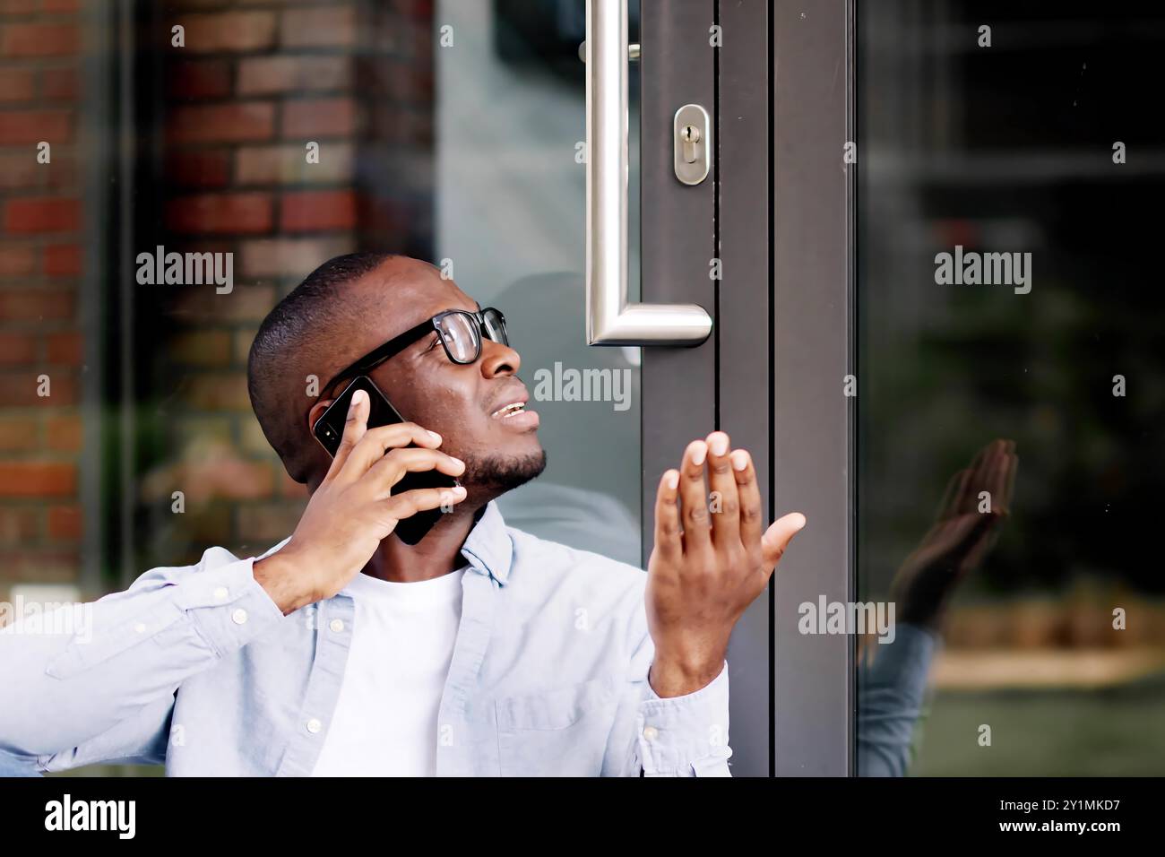 Frustrato uomo afroamericano chiuso fuori casa, aspetta Locksmith, utilizza l'app smartphone per l'assistenza di accesso. Foto Stock
