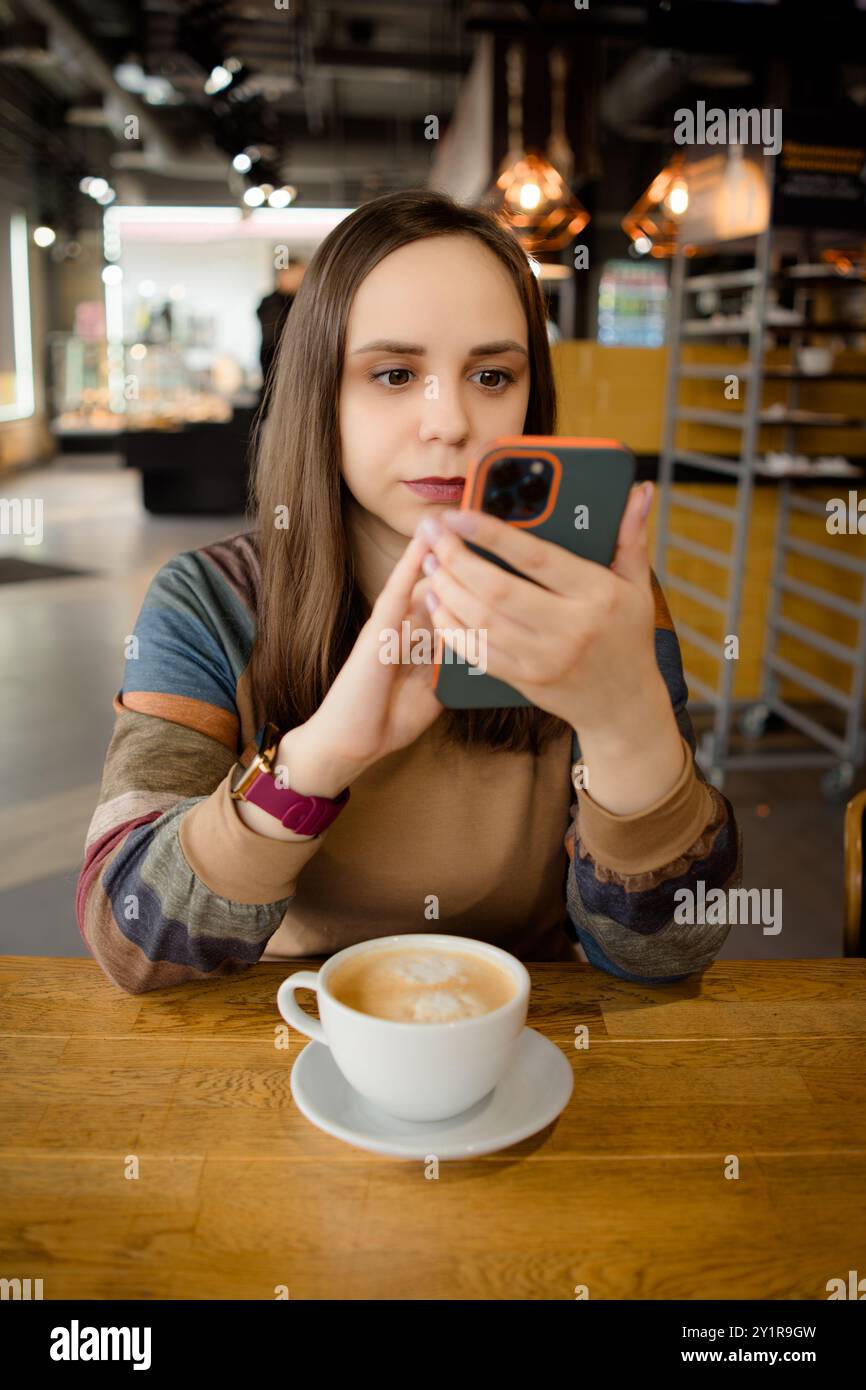 Una donna sfoglia intensamente il suo telefono, una tazza di caffè nelle vicinanze, nell'atmosfera invitante di un caffè locale Foto Stock