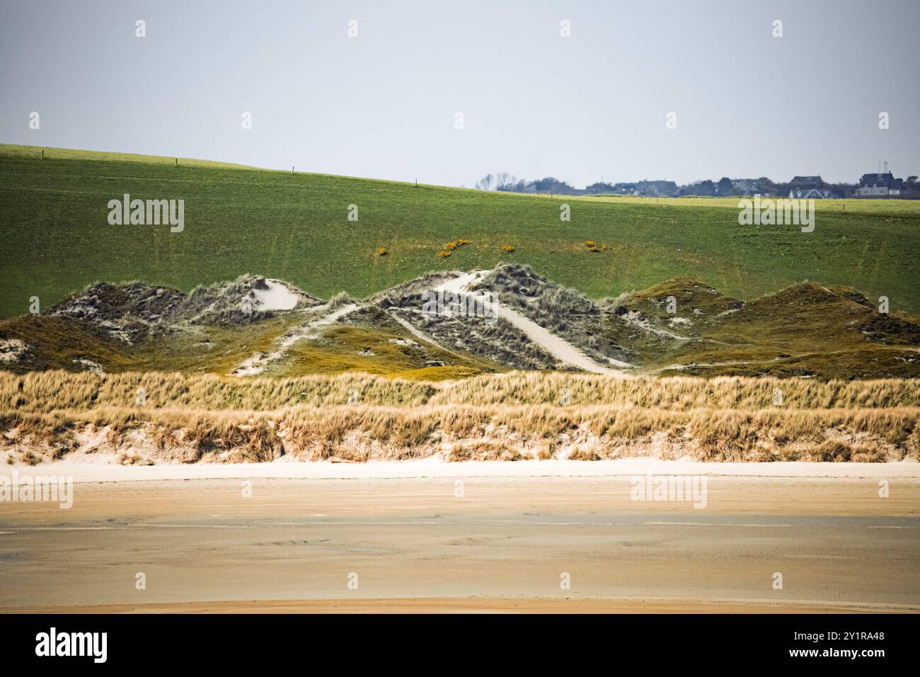 La spiaggia sabbiosa presenta dune naturali con bassa vegetazione e uno sfondo di colline verdi ondulate sotto un cielo limpido di giorno, creando una coasta pacifica Foto Stock