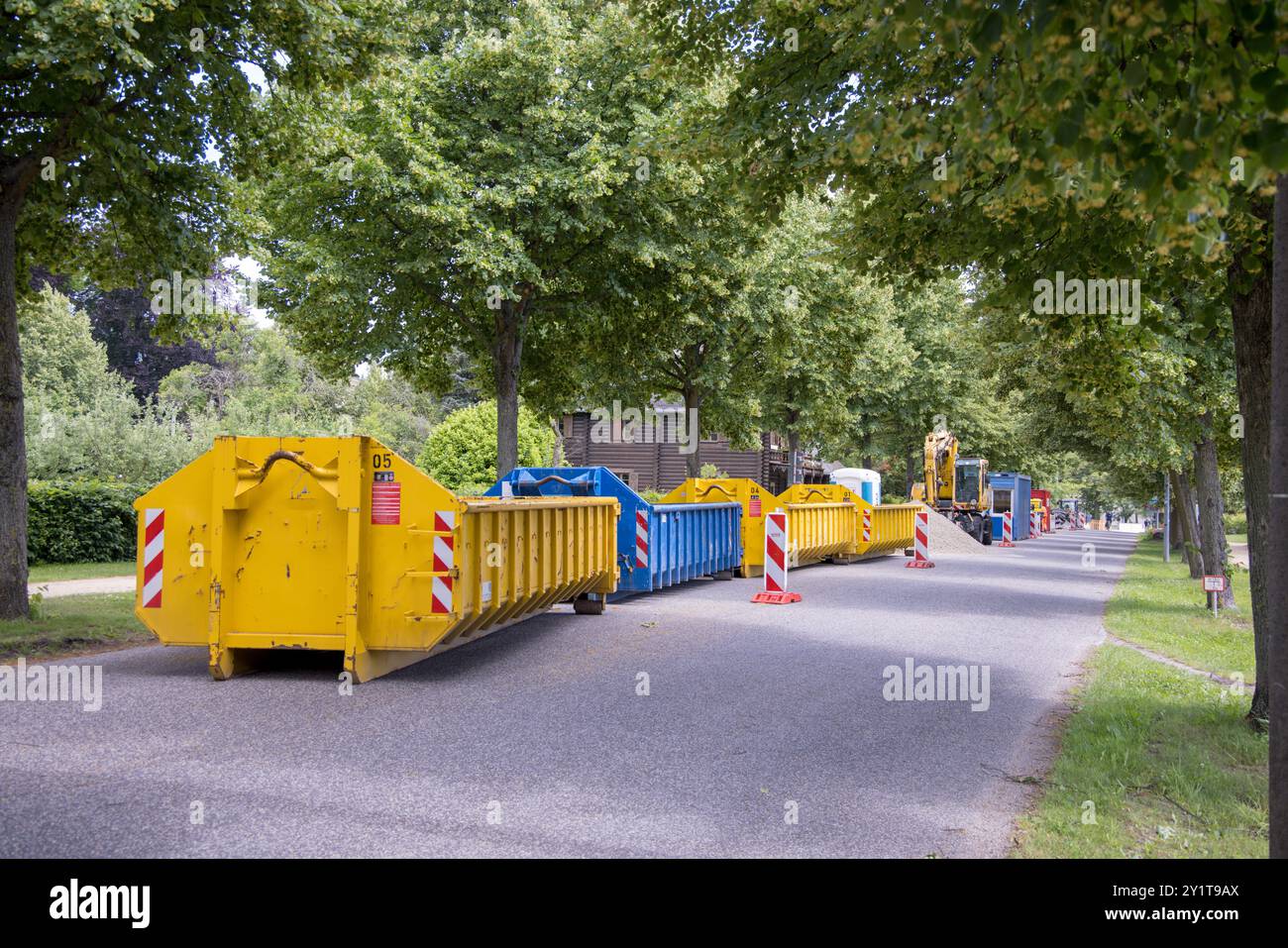 Contenitori gialli e bianchi per rifiuti da costruzione (cassonetti) parcheggiati su una strada con alberi verdi a Potsdam, Brandeburgo, Germania; lavori stradali Foto Stock