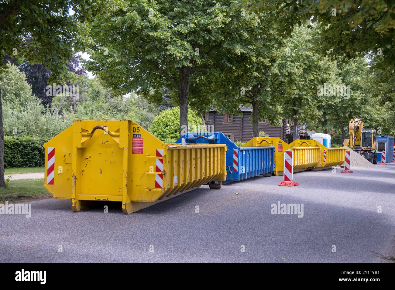Contenitori gialli e bianchi per rifiuti da costruzione (cassonetti) parcheggiati su una strada con alberi verdi a Potsdam, Brandeburgo, Germania; lavori stradali Foto Stock