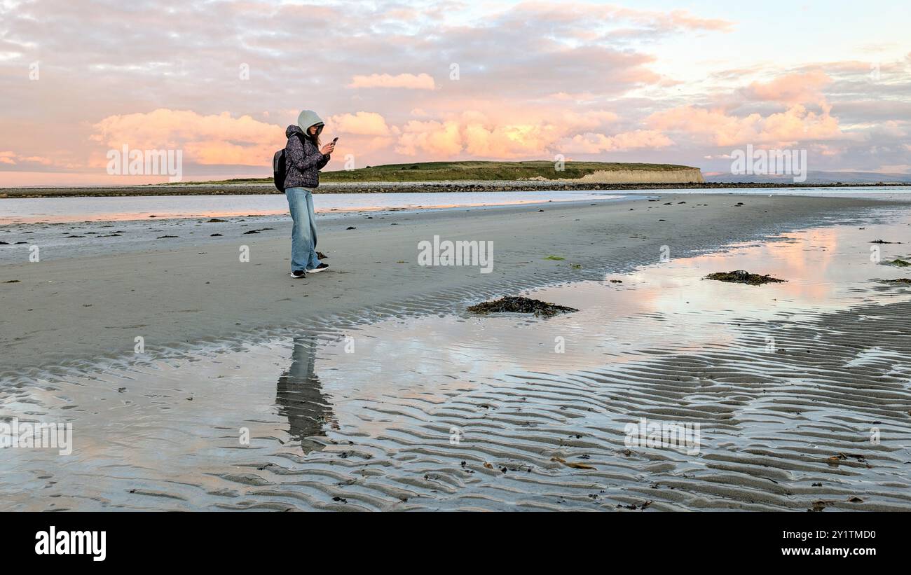 Donna escursionista con zaino in spalla che gode del paesaggio costiero del tramonto sulla spiaggia sabbiosa di Silverstrand, Galway, Irlanda, avventura e stile di vita, persone nella natura Foto Stock
