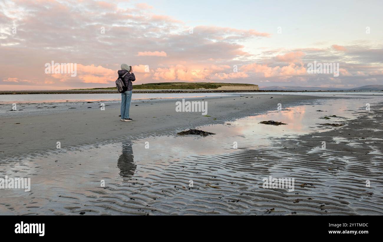 Donna escursionista con zaino in spalla che gode del paesaggio costiero del tramonto sulla spiaggia sabbiosa di Silverstrand, Galway, Irlanda, avventura e stile di vita, persone nella natura Foto Stock