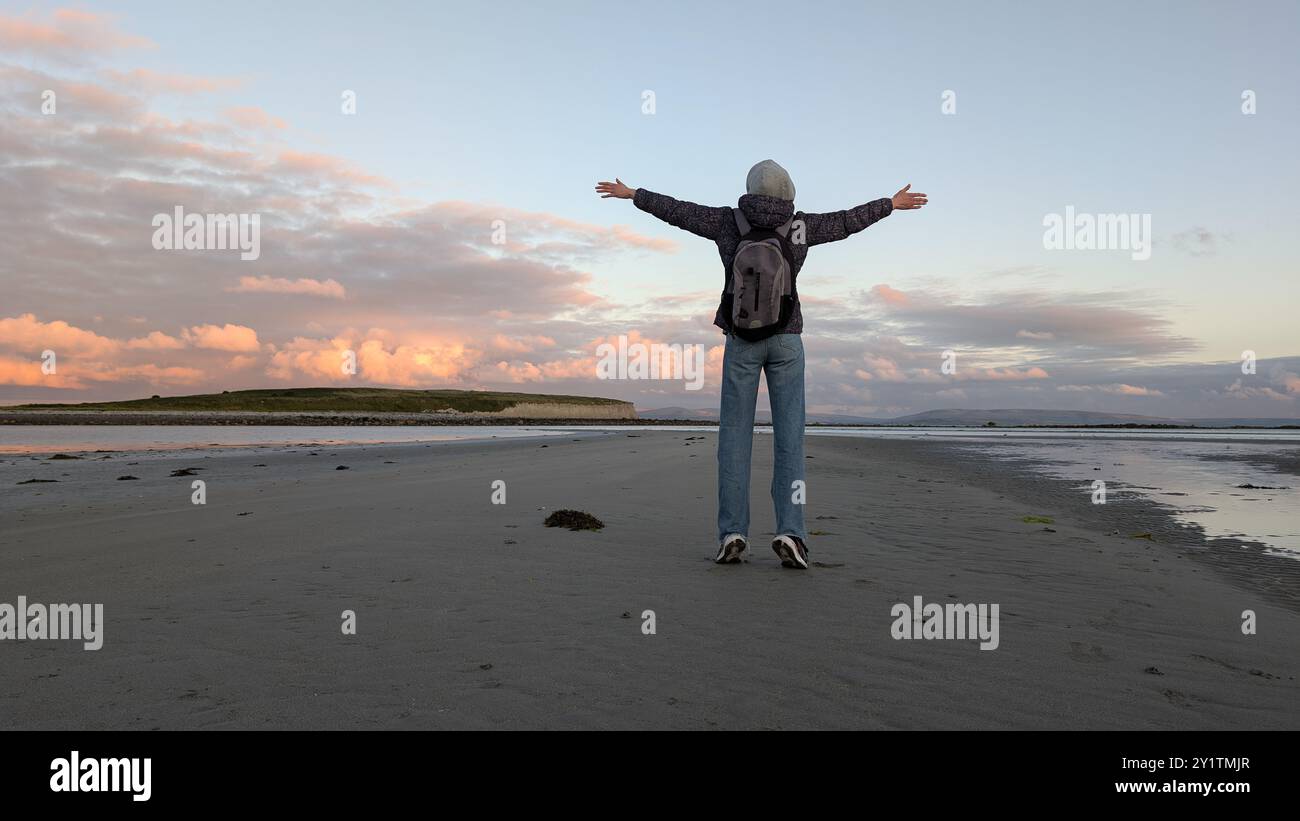 Donna escursionista con zaino in spalla che gode del paesaggio costiero del tramonto sulla spiaggia sabbiosa di Silverstrand, Galway, Irlanda, avventura e stile di vita, persone nella natura Foto Stock
