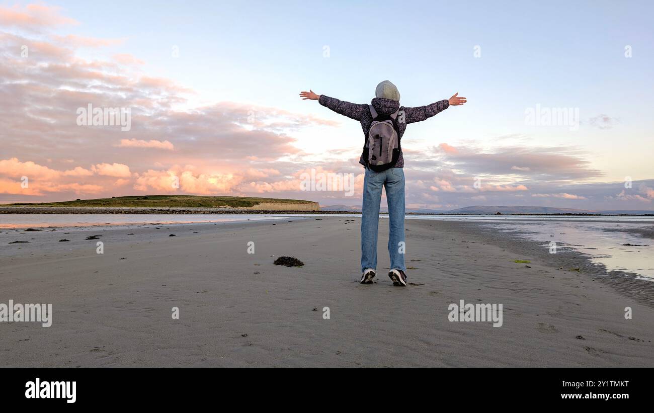 Donna escursionista con zaino in spalla che gode del paesaggio costiero del tramonto sulla spiaggia sabbiosa di Silverstrand, Galway, Irlanda, avventura e stile di vita, persone nella natura Foto Stock