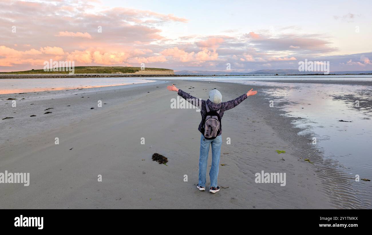 Donna escursionista con zaino in spalla che gode del paesaggio costiero del tramonto sulla spiaggia sabbiosa di Silverstrand, Galway, Irlanda, avventura e stile di vita, persone nella natura Foto Stock