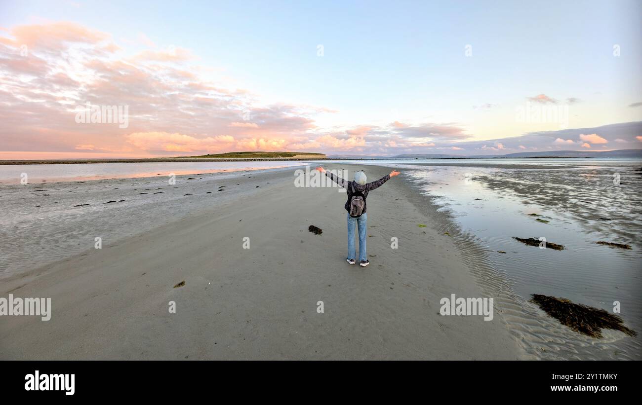 Donna escursionista con zaino in spalla che gode del paesaggio costiero del tramonto sulla spiaggia sabbiosa di Silverstrand, Galway, Irlanda, avventura e stile di vita, persone nella natura Foto Stock