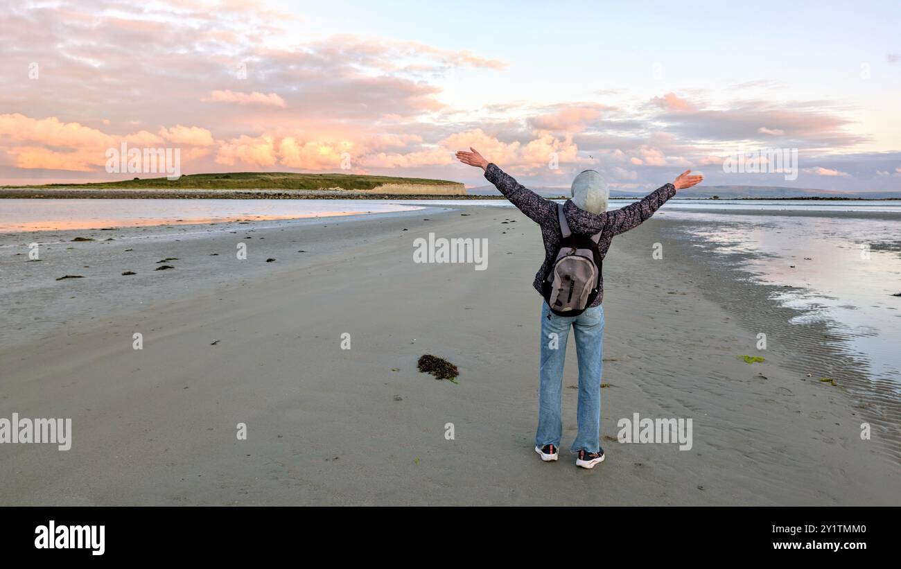 Donna escursionista con zaino in spalla che gode del paesaggio costiero del tramonto sulla spiaggia sabbiosa di Silverstrand, Galway, Irlanda, avventura e stile di vita, persone nella natura Foto Stock