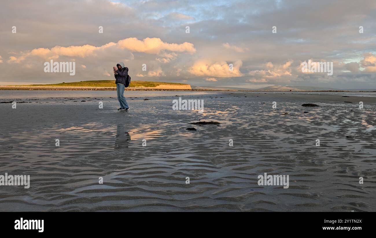 Donna escursionista con zaino in spalla che gode del paesaggio costiero del tramonto sulla spiaggia sabbiosa di Silverstrand, Galway, Irlanda, avventura e stile di vita, persone nella natura Foto Stock