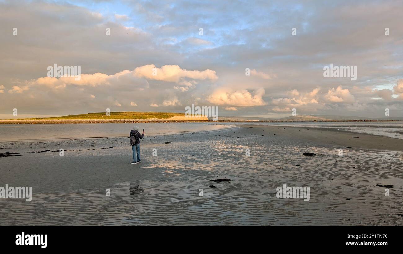 Donna escursionista con zaino in spalla che gode del paesaggio costiero del tramonto sulla spiaggia sabbiosa di Silverstrand, Galway, Irlanda, avventura e stile di vita, persone nella natura Foto Stock