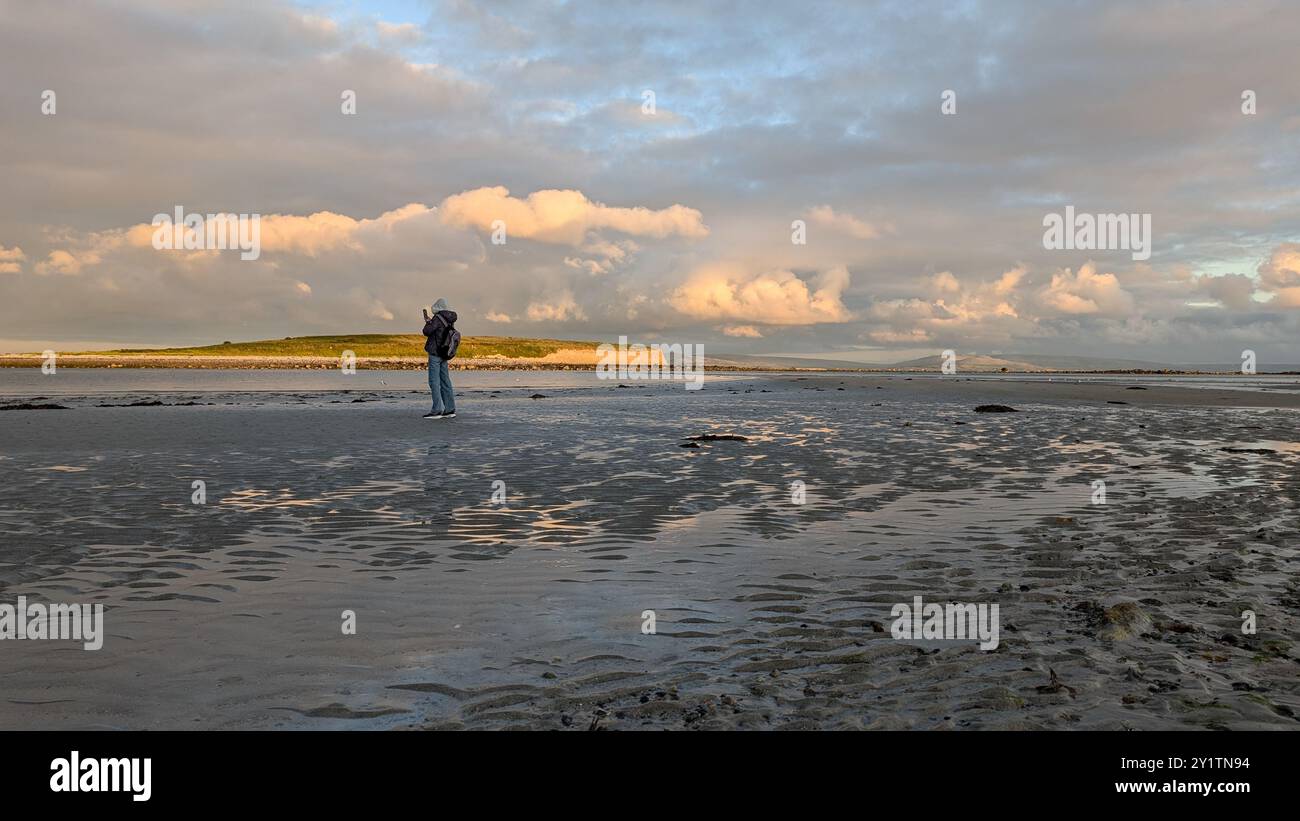Donna escursionista con zaino in spalla che gode del paesaggio costiero del tramonto sulla spiaggia sabbiosa di Silverstrand, Galway, Irlanda, avventura e stile di vita, persone nella natura Foto Stock