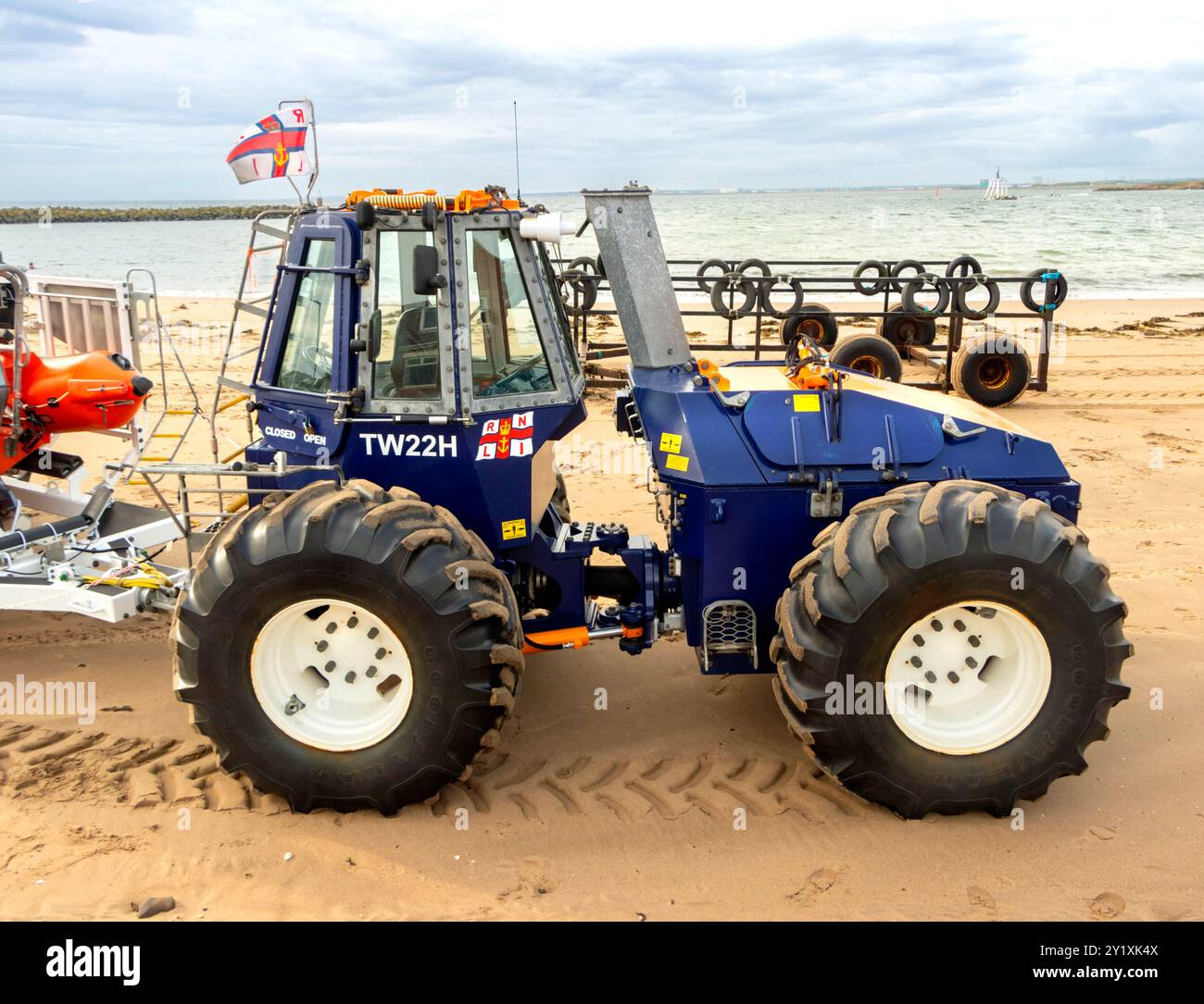 Un trattore di soccorso blu per impieghi gravosi RNLI parcheggiato su una spiaggia sabbiosa vicino all'oceano con una bandiera in cima e un rimorchio per scialuppa di salvataggio attaccato. Foto Stock