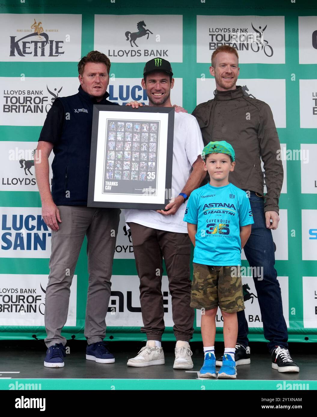 Mark Cavendish (centro) viene presentato con un collage fotografico del suo record di 35 vittorie al Tour De France - presentato da ed Clancy OBE (a sinistra) dopo la sesta tappa del Lloyds Bank Tour of Britain 2024 da Lowestoft a Felixstowe. Data foto: Domenica 8 settembre 2024. Foto Stock