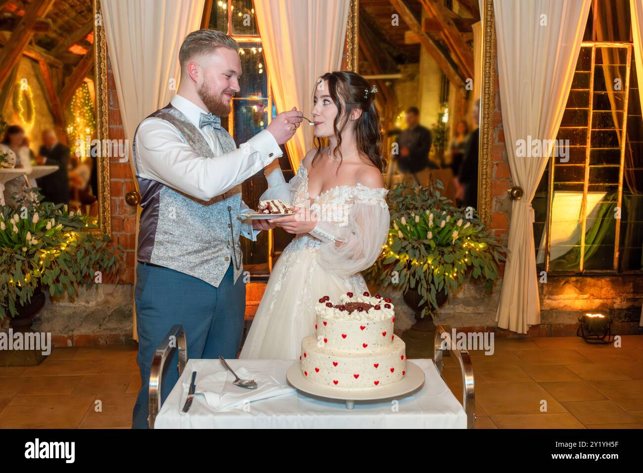 Uno sposo dà da mangiare alla sua sposa una fetta di gateau della Foresta Nera come torta nuziale durante il loro ricevimento di nozze. La coppia sorride in un ambiente caldo e romantico Foto Stock