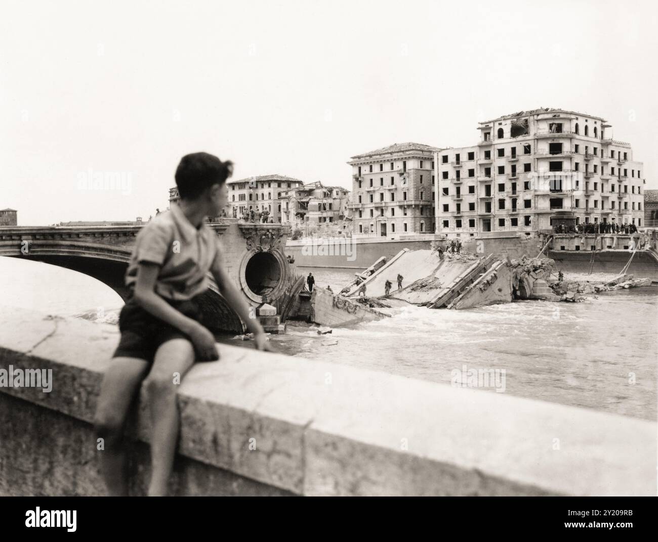 1945 , 26 aprile , VERONA , VENETO , ITALIA : DALLA DIDASCALIA ORIGINALE: »Quinta Armata americana a Verona . Il ragazzo italiano si affaccia sul fiume Adige e sul ponte della Vittoria che i tedeschi nazisti e fascisti italiani hanno fatto esplodere durante la fuga". La notte del 25 aprile 1945, quando fu estratta e fatta esplodere dai soldati tedeschi in ritirata, un'esplosione che lasciò intatta solo l'arco verso la città. In questa foto scattata da Lungadige Panvinio si può anche vedere la devastazione di tutte le case di fronte al ponte che si affaccia su Lungadige Giamomo Matteotti ( come oggi rinominato ) e Lungadige Camp Foto Stock