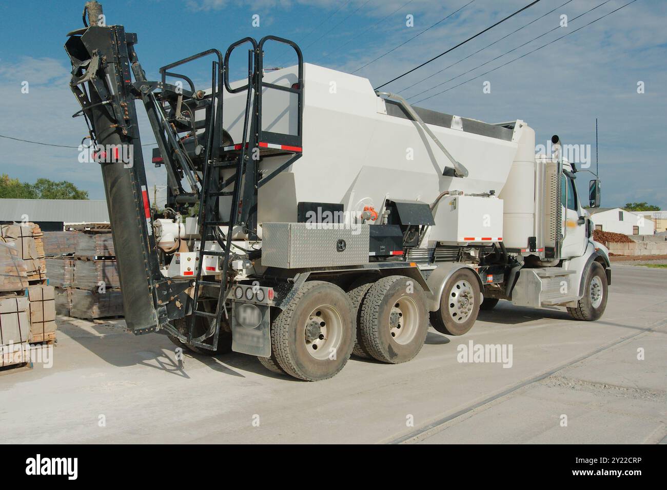 Vista laterale angolata di un grande camion di cemento bianco nel tardo pomeriggio con cielo blu e nuvole bianche sul retro. Binari ferroviari di fronte alla strada polverosa con edificio Foto Stock