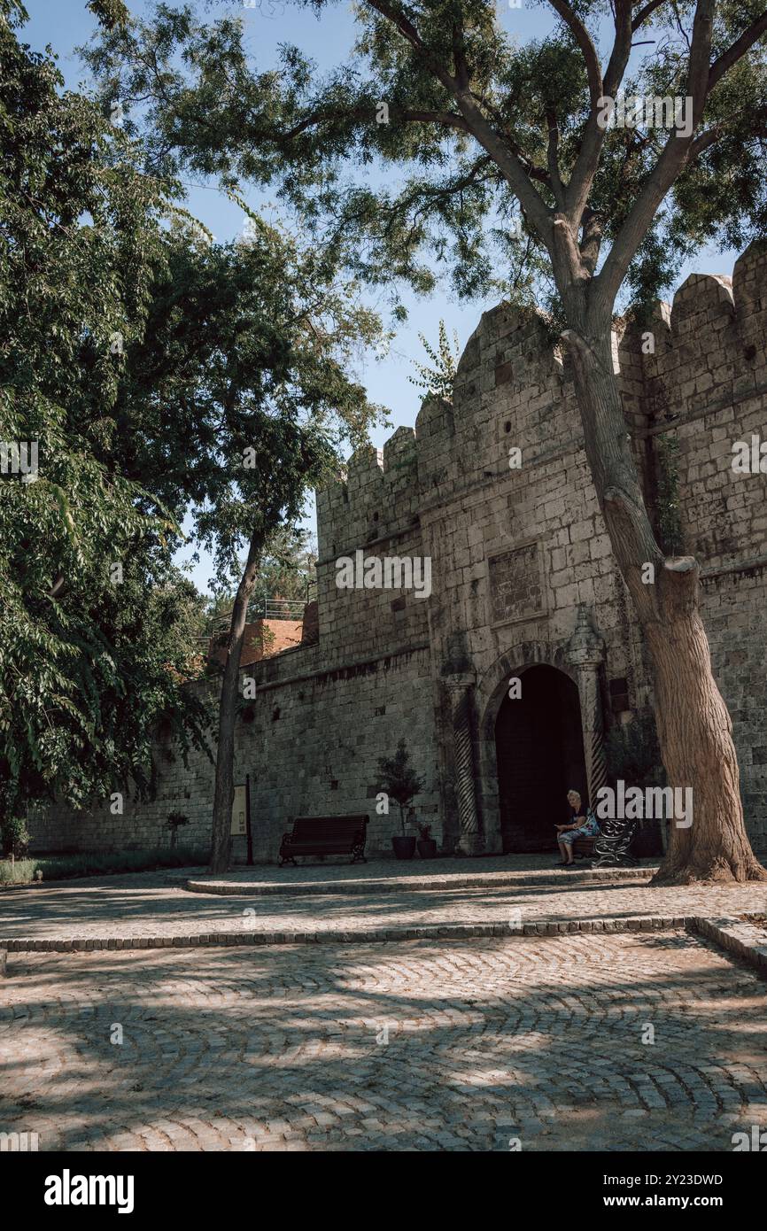 Una donna seduta all'ombra di alberi ad un ingresso laterale della Fortezza di Nis in Serbia in una giornata di sole, con vista sulle imponenti pareti. Foto Stock