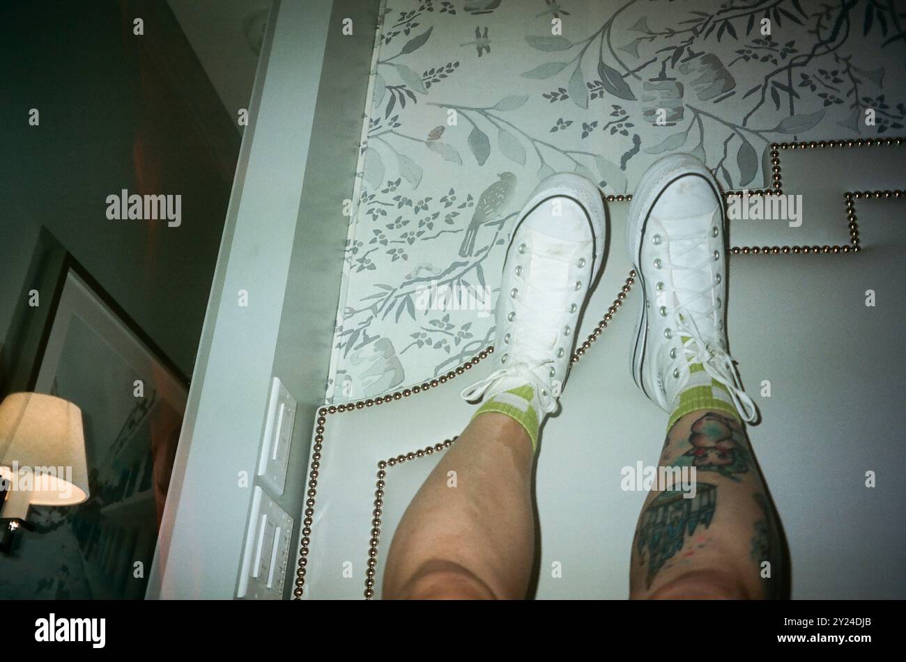 Gambe tatuate in sneakers bianche contro una parete decorativa, Foto Stock