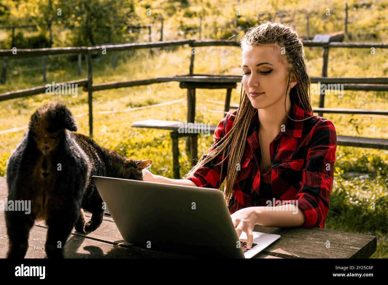 La giovane donna con lunghe trecce bionde è seduta a un tavolo di legno all'aperto con il suo gatto, lavorando a distanza sul suo laptop Foto Stock