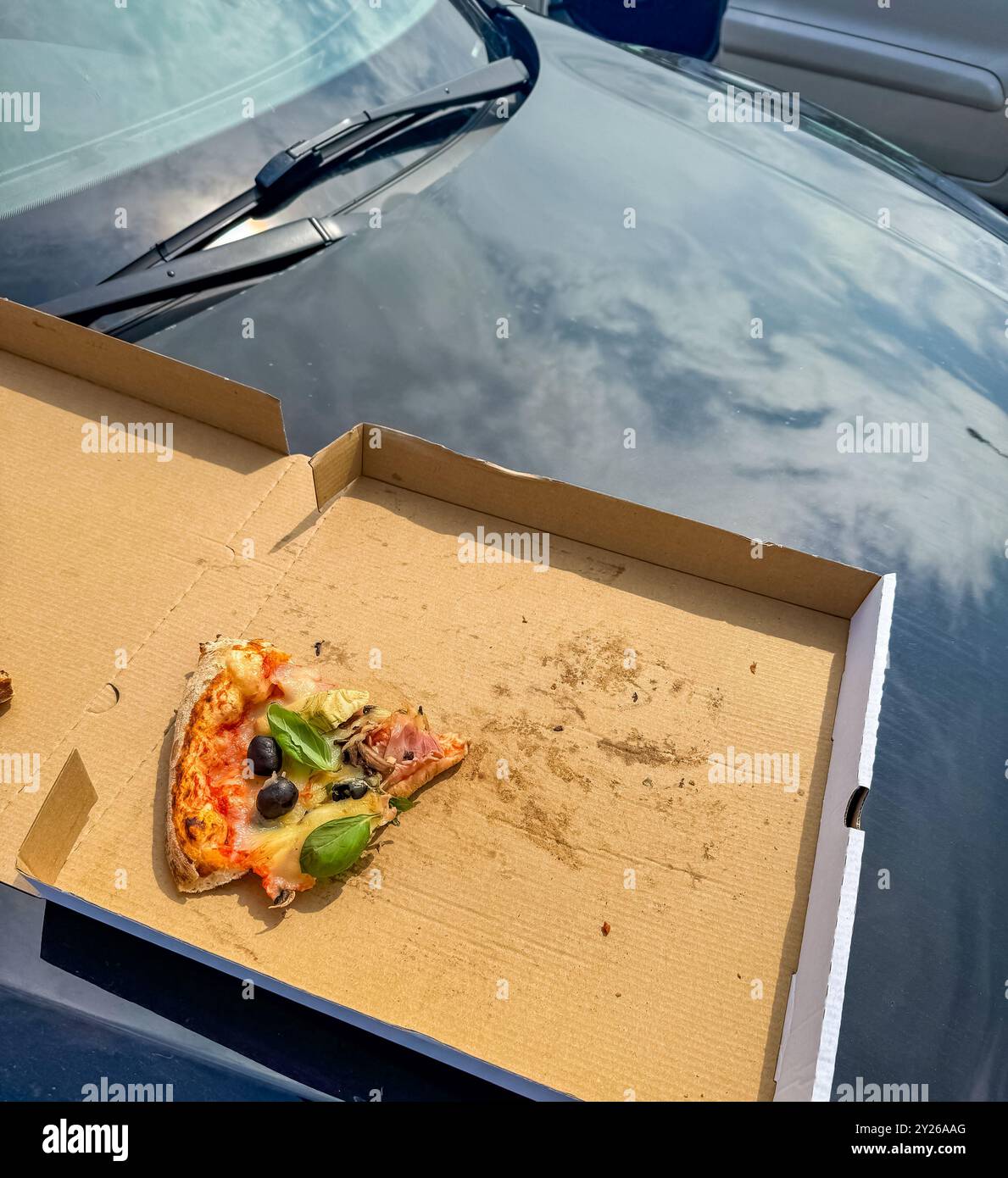 una scatola per pizza con una fetta di pizza all'interno, sul cofano di un'auto nera. Foto di alta qualità Foto Stock