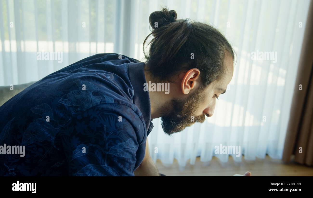 Un uomo con la barba siede in una stanza luminosa, guardando attentamente il suo smartphone mentre riposa la mano su un libro aperto. Foto Stock