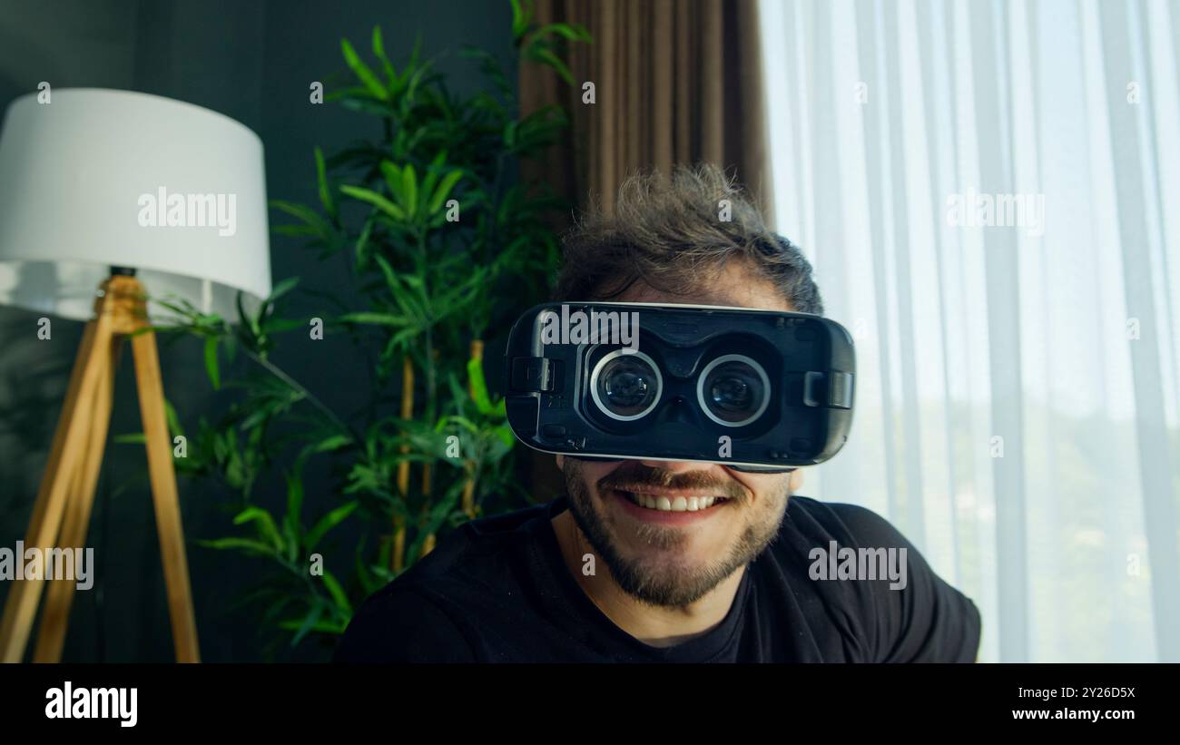 Un uomo divertente sorride mentre usa un visore VR in un salotto moderno Foto Stock