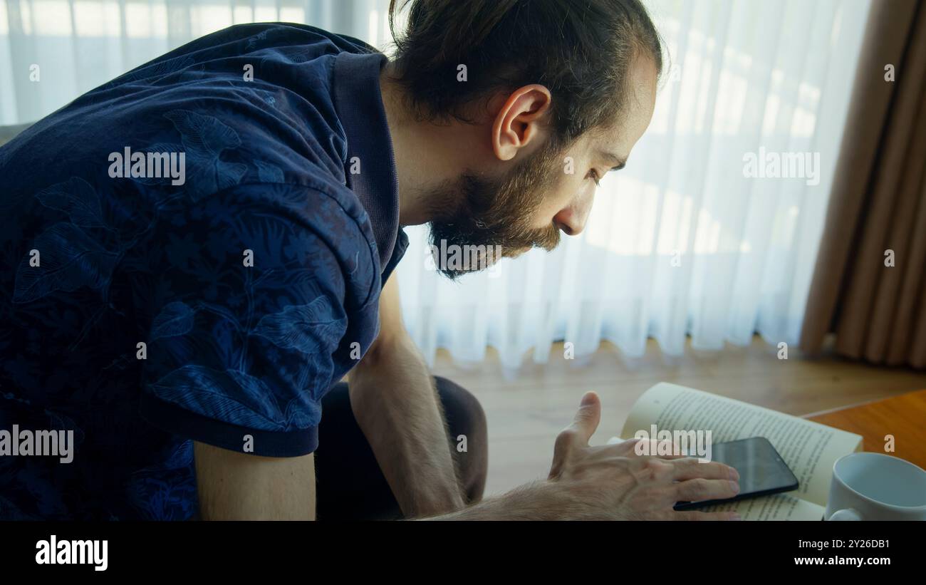 Un uomo con la barba siede in una stanza luminosa, guardando attentamente il suo smartphone. Foto Stock