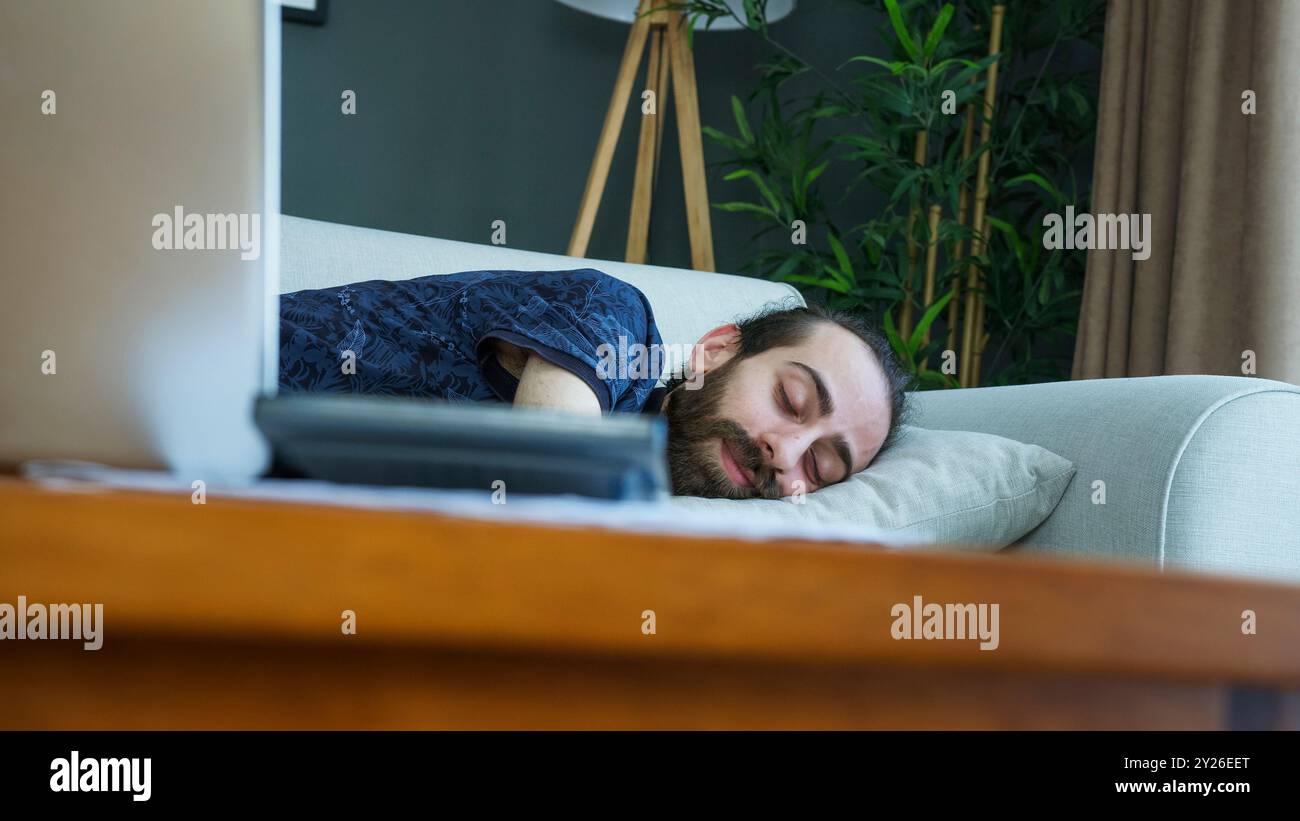 Un uomo sta riposando su un divano, dopo essersi addormentato davanti al suo portatile. Stanchezza o relax dopo una lunga giornata di lavoro o di tempo sullo schermo a casa. Foto Stock