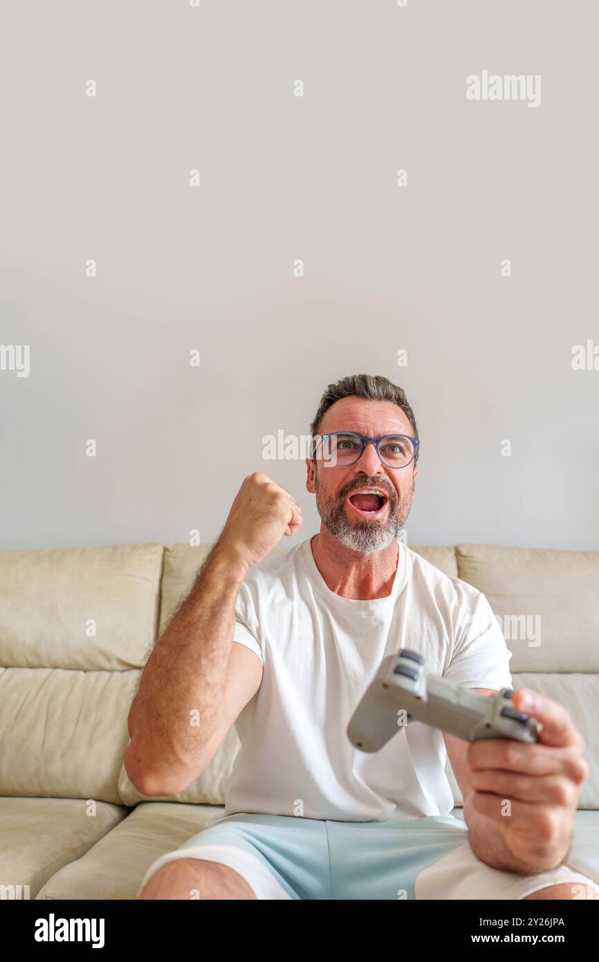 Vista frontale dell'uomo di mezza età che celebra la vittoria mentre gioca ai videogiochi sul divano a casa Foto Stock