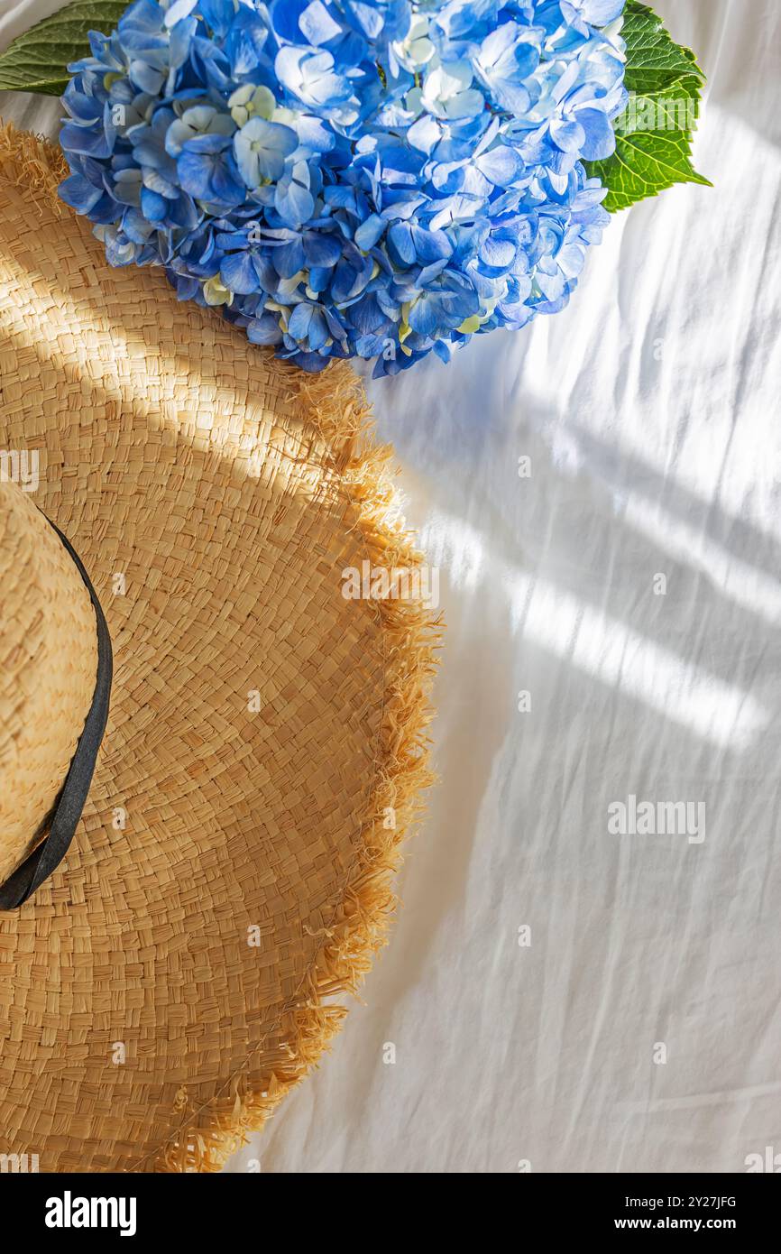 Un cappello di paglia e fiori di ortensia blu su un copriletto bianco con soffice luce mattutina Foto Stock