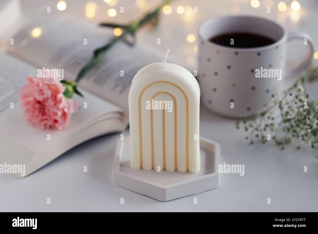 Candela bianca su un tavolo con un libro, tazza e fiori sullo sfondo. Arredi per la casa. Interni accoglienti. Foto Stock