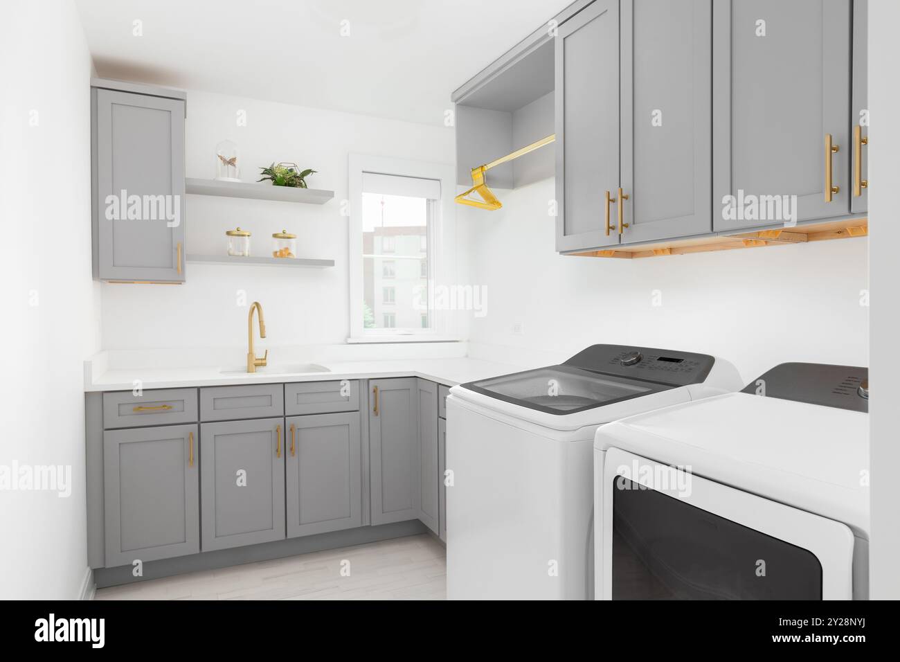 Una lavanderia con armadi grigi, lavatrice e asciugatrice bianca, pavimento piastrellato, piano di appoggio bianco, rubinetto e ferramenta dorati. Nessun marchio o laboratorio Foto Stock