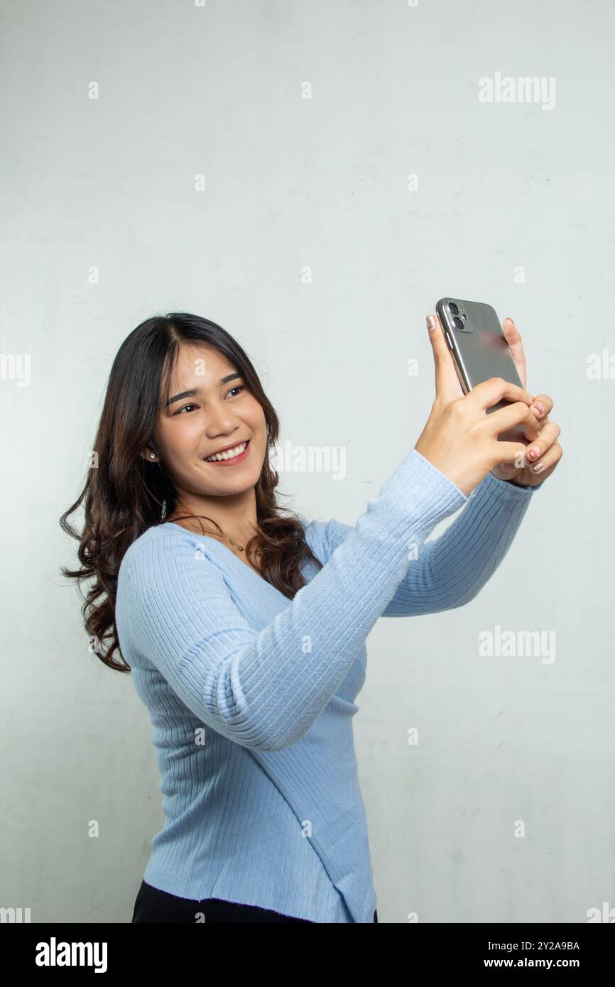 Bella ragazza asiatica in tuta casual azzurra che scatta selfie con il telefono cellulare e sorride felicemente, donna adulta in piedi uso nella pubblicità Foto Stock