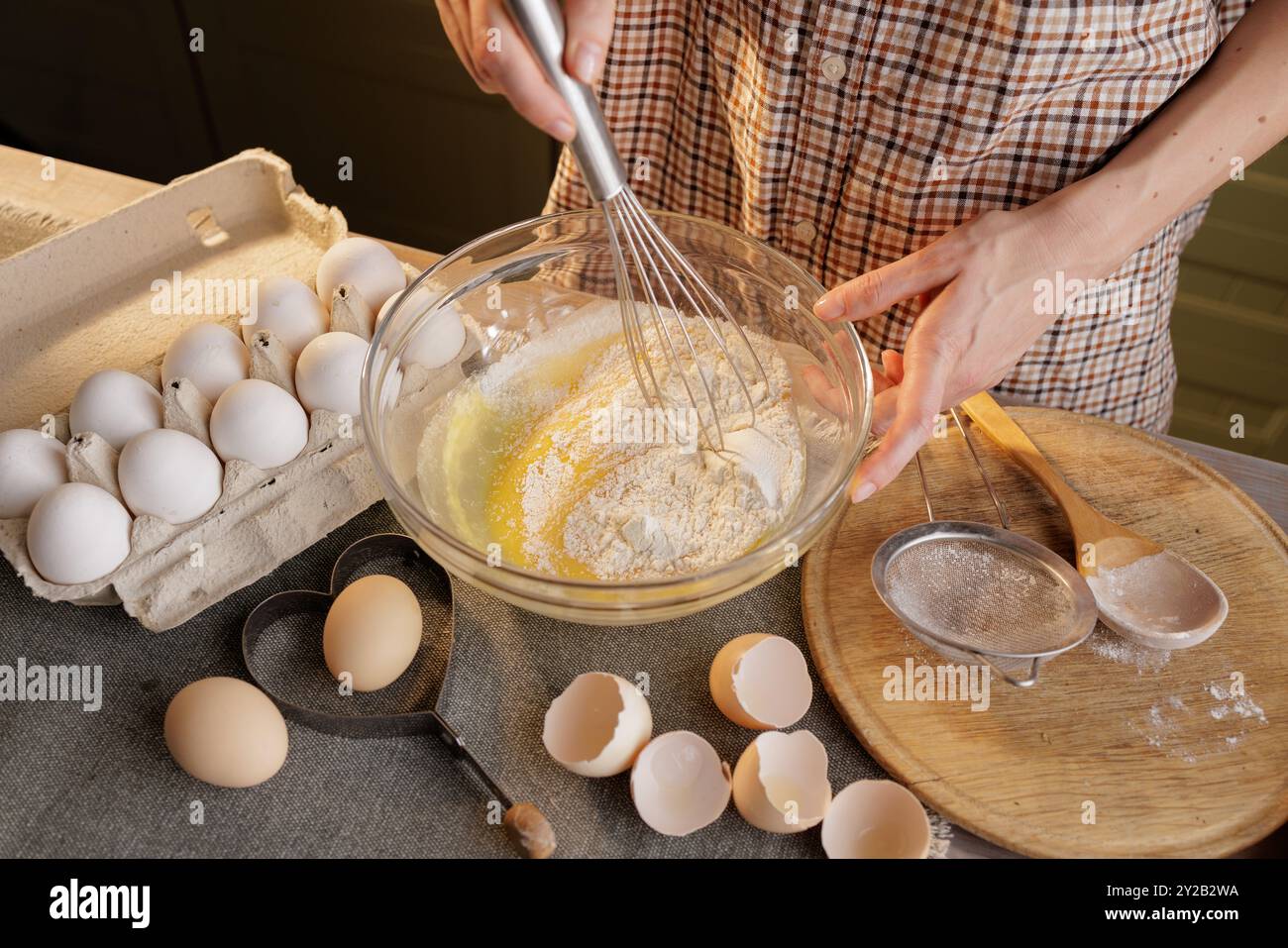 Primo piano di una donna che prepara l'impasto in un recipiente sullo sfondo della cucina di casa, vassoio di uova, gusci d'uovo e farina sul tavolo Foto Stock
