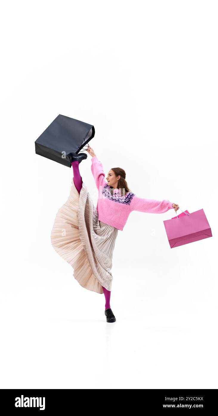 Vestita con una gonna a pieghe fluide e un pullover rosa, la donna prende il sopravvento tenendo in mano le borse dello shopping su sfondo bianco. Foto Stock