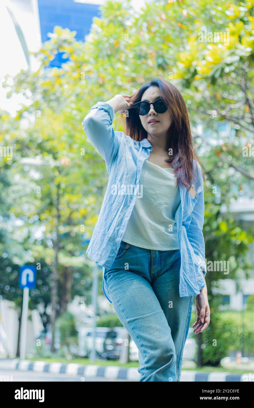 Elegante donna asiatica con occhiali da sole che si pettina i capelli in piedi sul percorso all'aperto del parco. studentesse per i temi dell'educazione, della moda e dello stile di vita Foto Stock