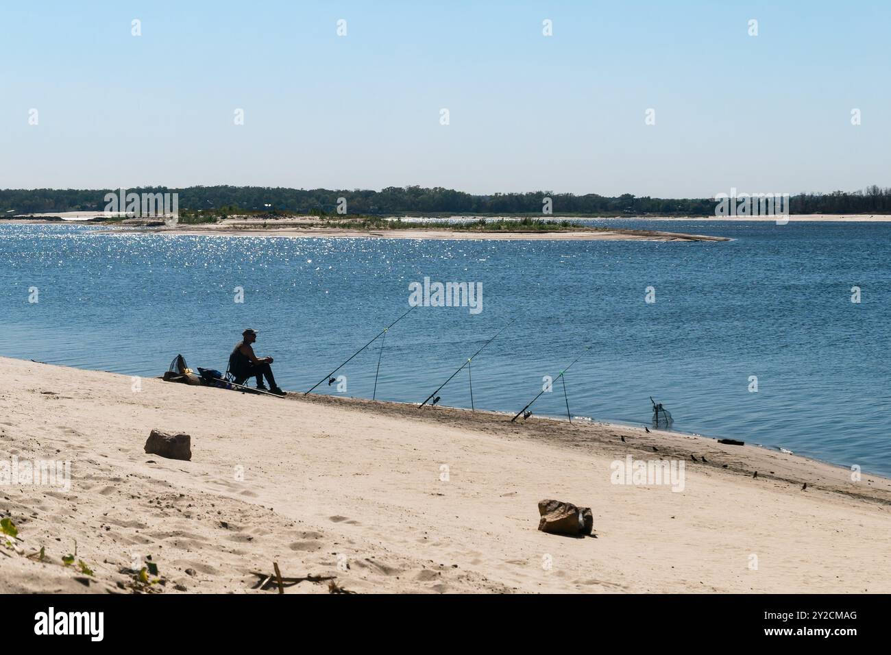 Zaporizhzhia, Ucraina - 7 settembre 2024: Un pescatore solitario con diverse canne da pesca siede sulla riva sabbiosa di un ampio fiume in una giornata di sole Foto Stock Zaporizhzhia, Ucraina - 7 settembre 2024: Un pescatore solitario con diverse canne da pesca siede sulla riva sabbiosa di un ampio fiume in una giornata di sole Foto Stock