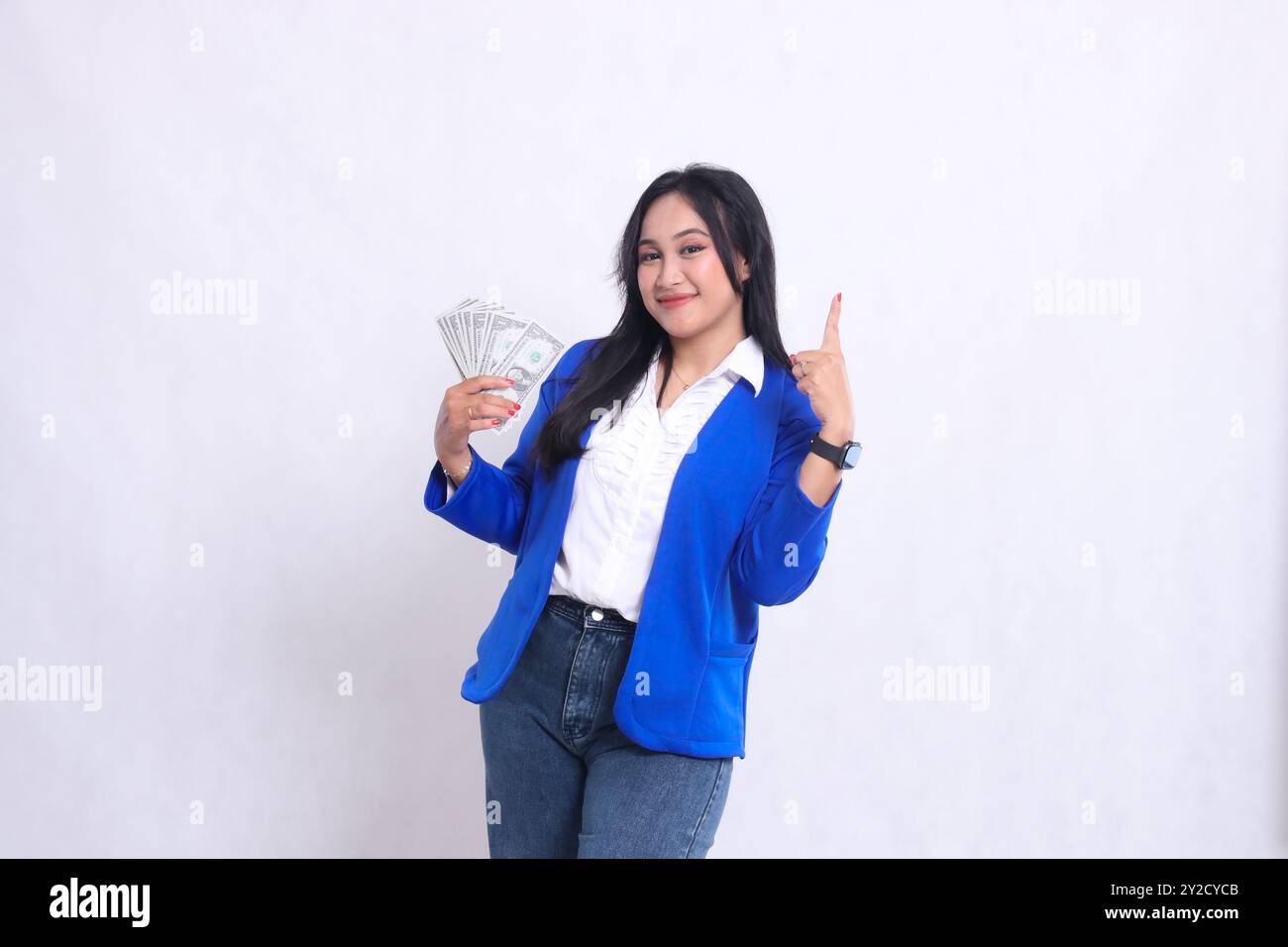 bella ragazza d'ufficio adulta che indossa una tuta formale blu in piedi sorridente alla macchina fotografica che tiene il conto del dollaro a destra e punta su sfondo bianco isolato Foto Stock