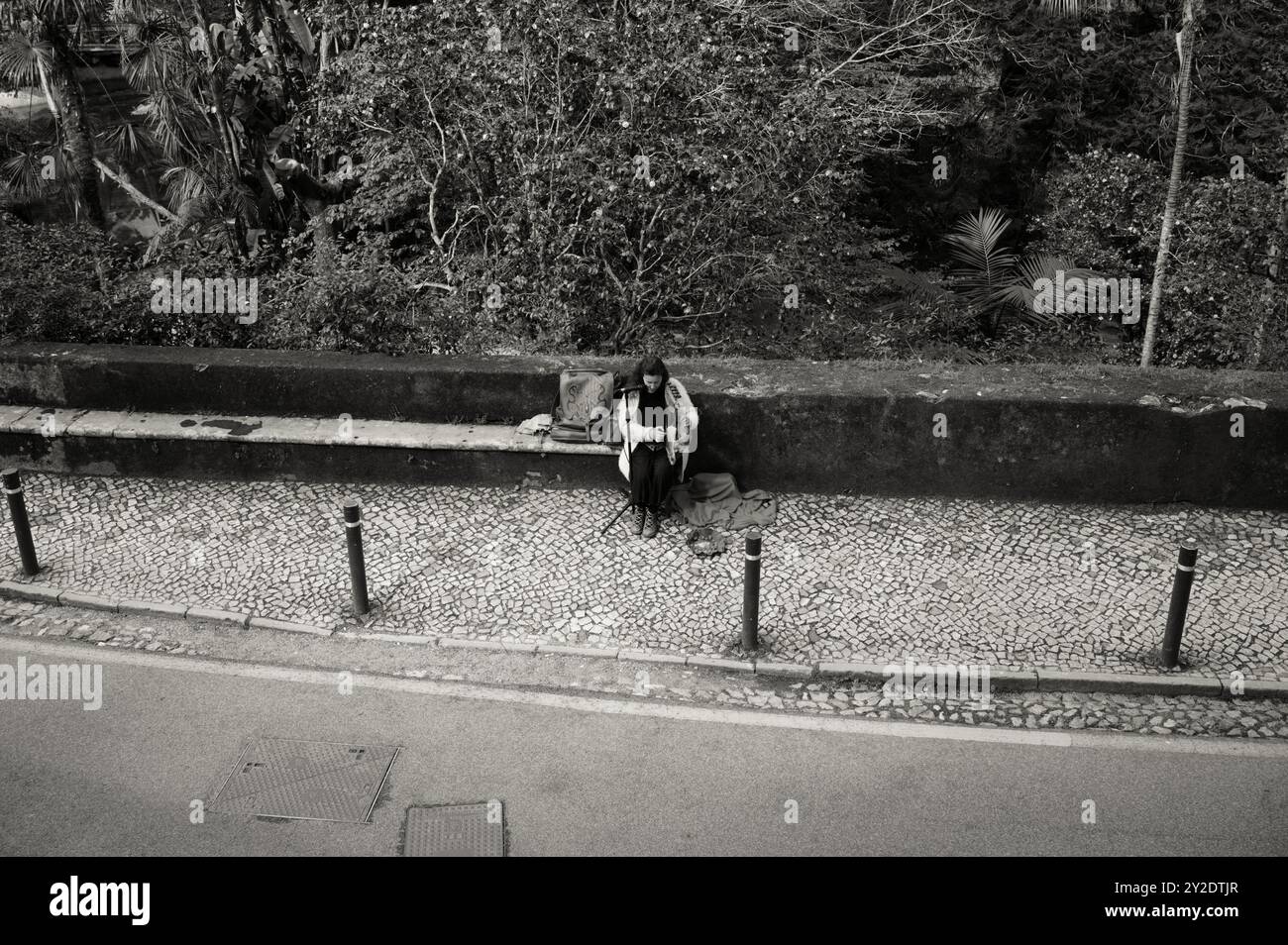 Monocromatico di una musicista di strada seduta su un sentiero acciottolato, che suona il suo strumento simile a un'arpa a Sintra Foto Stock