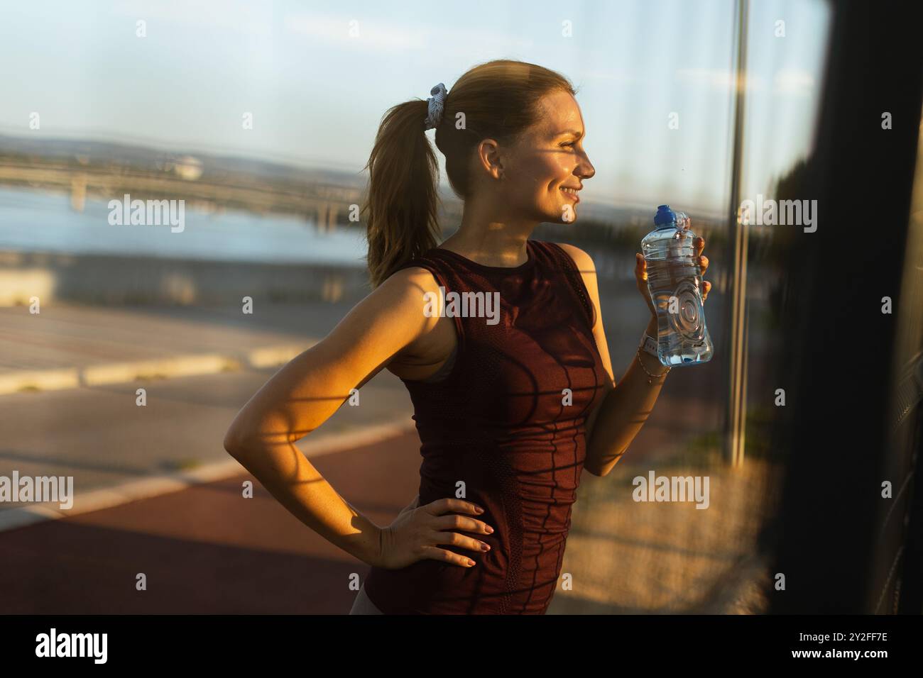 Con il sole che tramonta alle sue spalle, la donna si idrata dopo un allenamento all'aperto rinvigorente, prendendosi un momento per apprezzare la serena atmosfera lungo il fiume Foto Stock