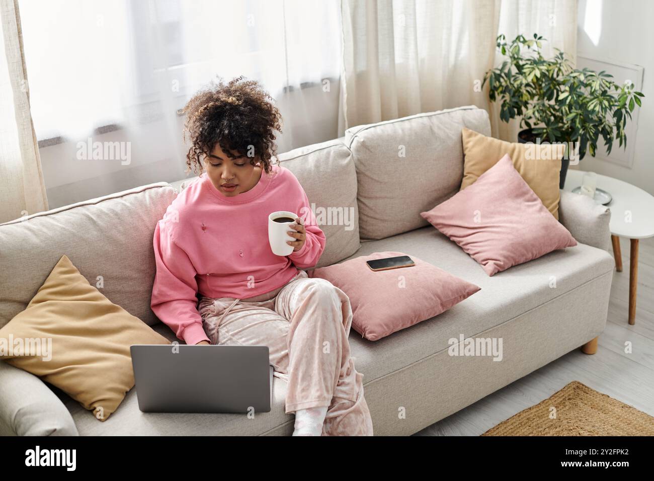 Una donna si rilassa all'interno, lavorando sul suo notebook con una bevanda calda in mano. Foto Stock