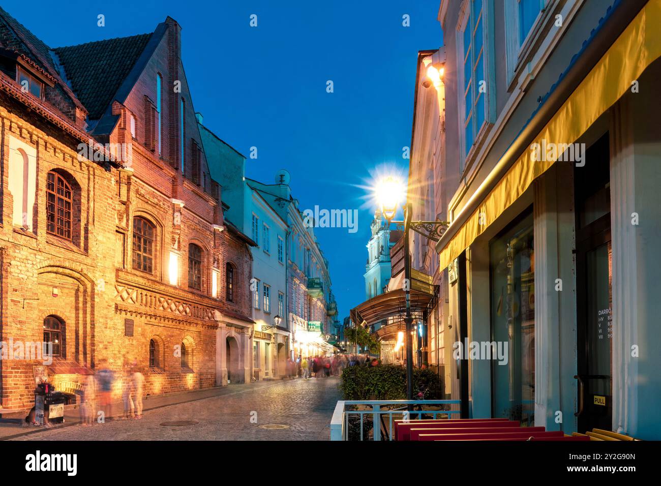Strada acciottolata della città vecchia illuminata da caldi lampioni di strada la sera. Edifici storici, caffetterie accoglienti e gente sfocata dal movimento. Freddo destin Foto Stock