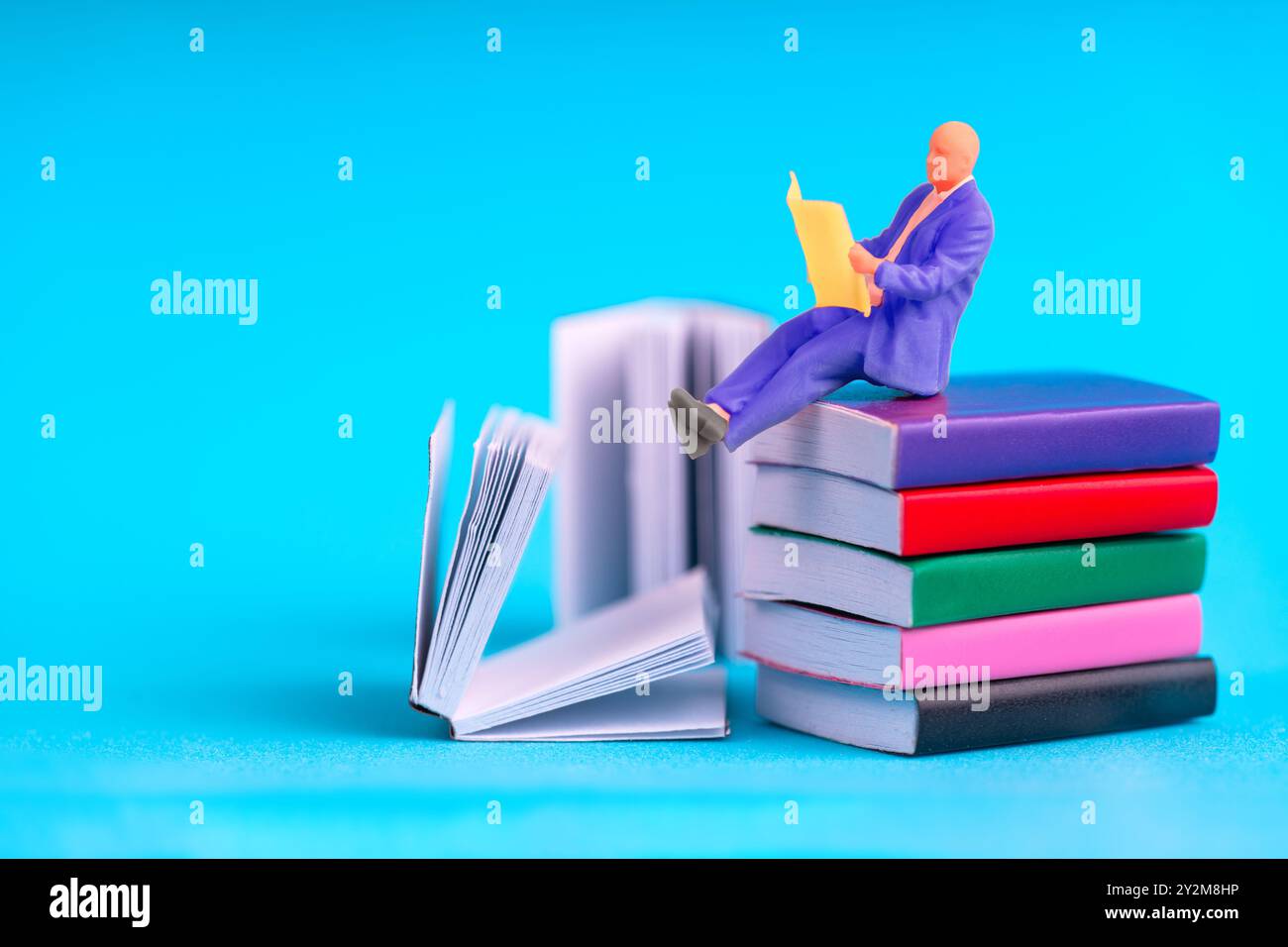 Concetto di apprendimento e conoscenza con un uomo in miniatura che indossa una tuta viola, si siede su libri vivaci e legge da una cartella aperta. Foto Stock