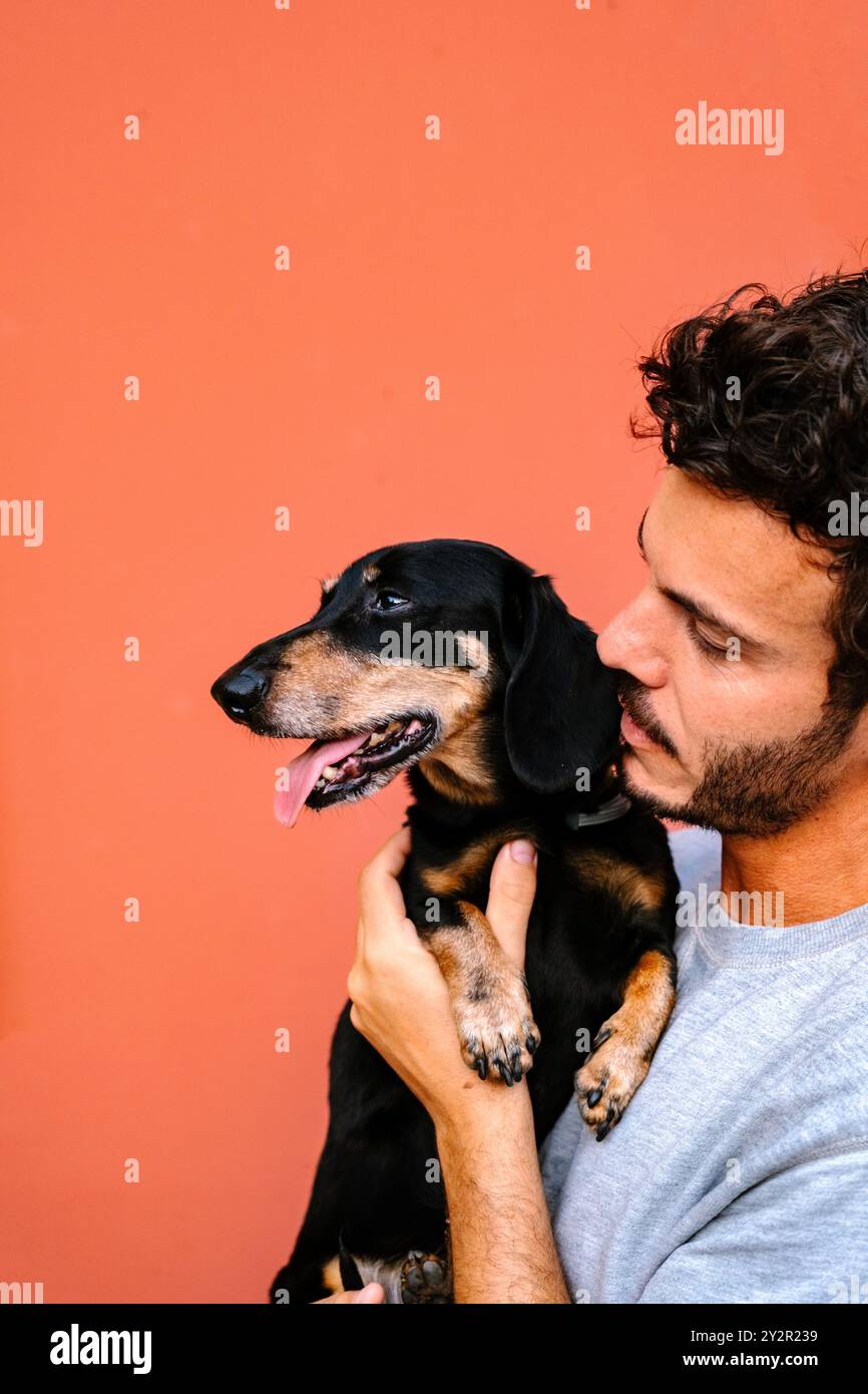 Un'immagine commovente che cattura un giovane maschio adulto affettuosamente che tiene e guarda il suo dachshund nero e abbronzante del cane, che sembra contento, mostra Foto Stock