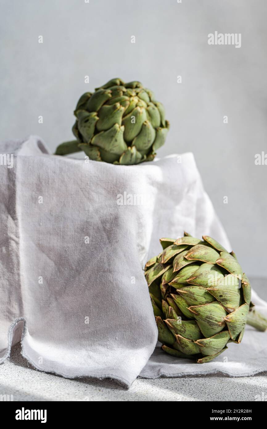 Due carciofi freschi e verdi poggiano su un panno di lino delicatamente drappeggiato, che mostra la loro texture dettagliata e naturale con uno sfondo morbido e neutro Foto Stock