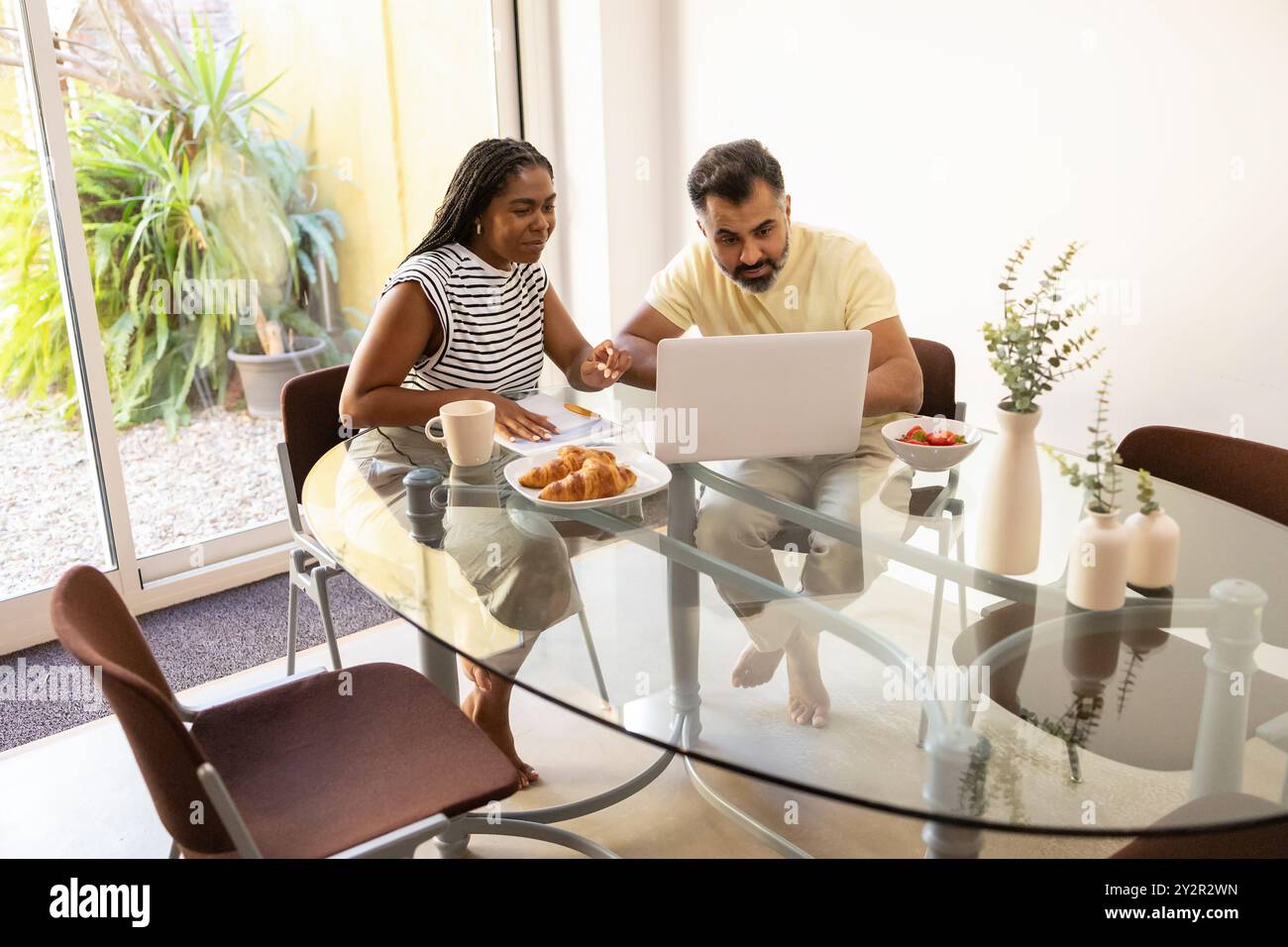 Una donna nera condivide un momento di gioia per la colazione con un uomo indiano mentre si impegna con un portatile in una luminosa e moderna sala da pranzo Foto Stock
