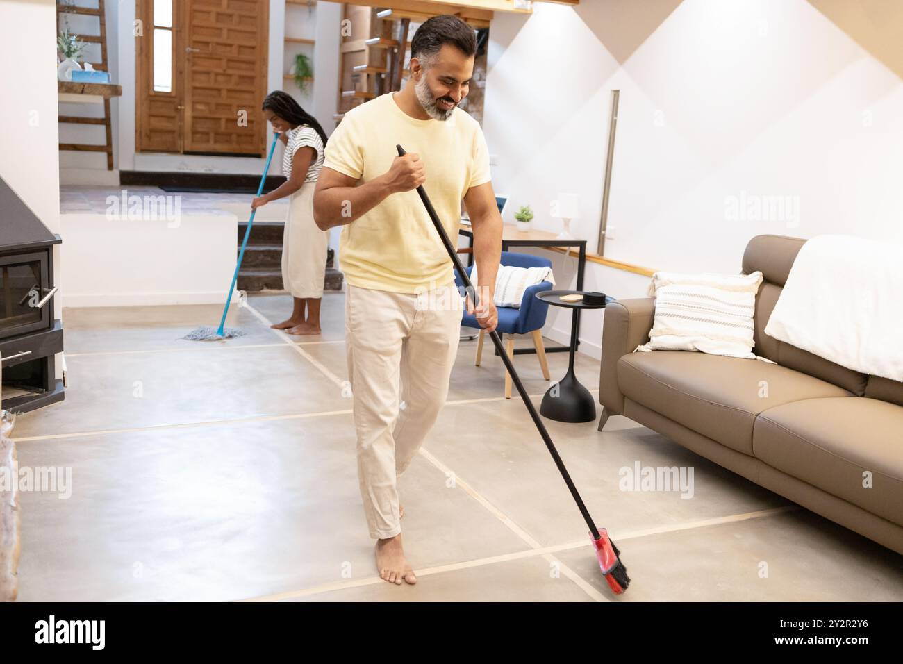 Un uomo indiano di mezza età e una donna nera spazzano via lo spazioso ed elegante soggiorno, mantenendo la loro casa ordinata e piacevole Foto Stock
