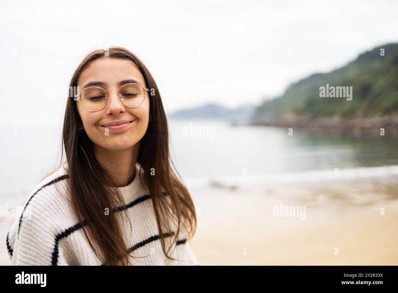 Giovane donna a occhi chiusi, sorridente contentosamente, si stende in riva al mare lungo la costa delle Asturie, Spagna, trasudando un'aura pacifica e riflessiva in mezzo a un Foto Stock